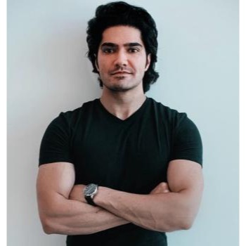 Portrait of a young man with dark hair, wearing a black t-shirt and a watch, arms crossed, standing against a light wall.