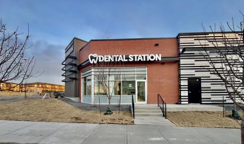 Front of a Dental Station clinic with a brick facade, glass doors, and a bold sign reading 'DENTAL STATION' above the entrance; bare trees and a sidewalk in the foreground.