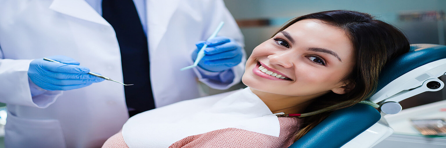 Smiling woman in a dental chair during a checkup, dentist in white coat and blue gloves holding dental tools beside her.