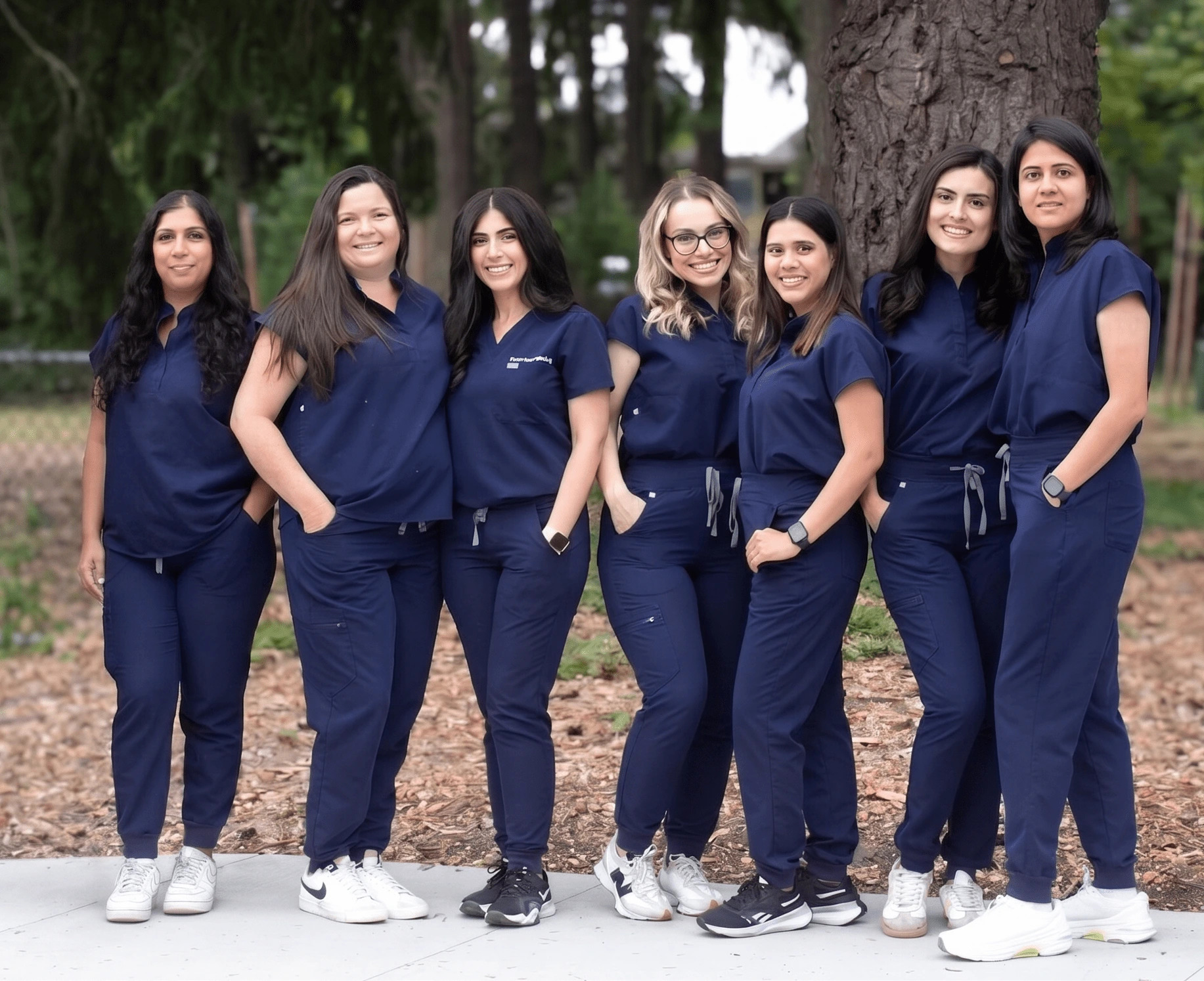 Group of seven women in navy scrubs posing outdoors, smiling and standing in a line by a tree in a park-like setting.