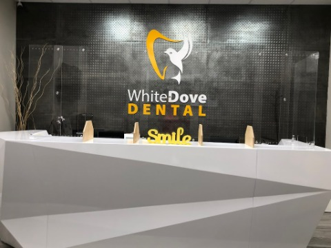 Reception area of White Dove Dental featuring a white angular desk, glass dividers, and a dark wall with the White Dove Dental logo and a 'Smile' sign.