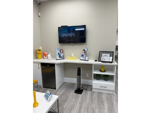 Reception counter in a modern office with a wall-mounted TV, framed photos, and a small fridge beneath the counter.