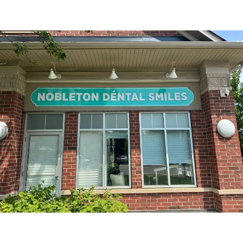 Exterior of a brick dental clinic with a teal sign that reads 'Nobleton Dental Smiles' above two large windows.