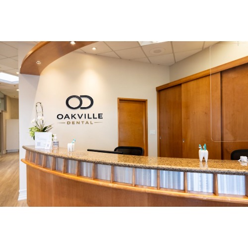 Reception desk at Oakville Dental with a curved wood counter, logo on the wall, and small plants on the counter.